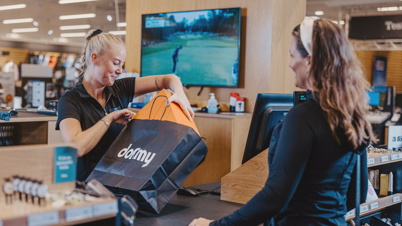 Butiksanställd räcker över en Dormy-shoppingpåse till en kund vid kassan i en sportbutik, med en TV som visar golf i bakgrunden.