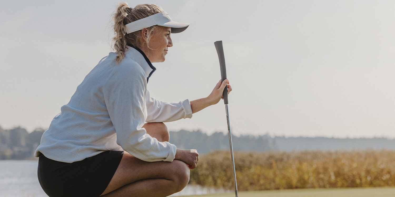 A woman in a white top and visor is crouching and holding a golf club on a golf course by the water.