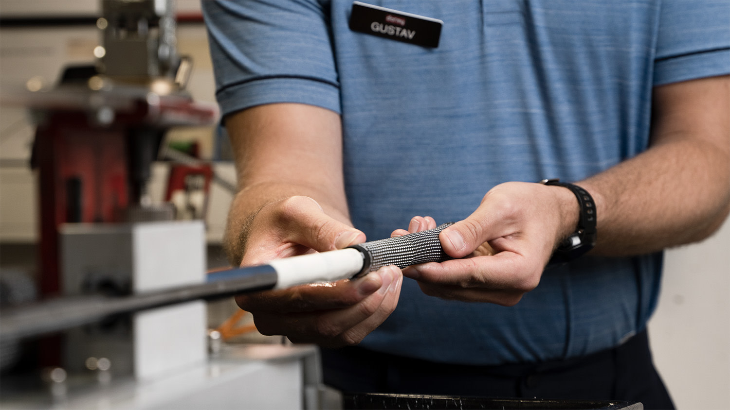 En person med namnskylt "Gustav" byter grepp på en golfklubba i en verkstadsmiljö.