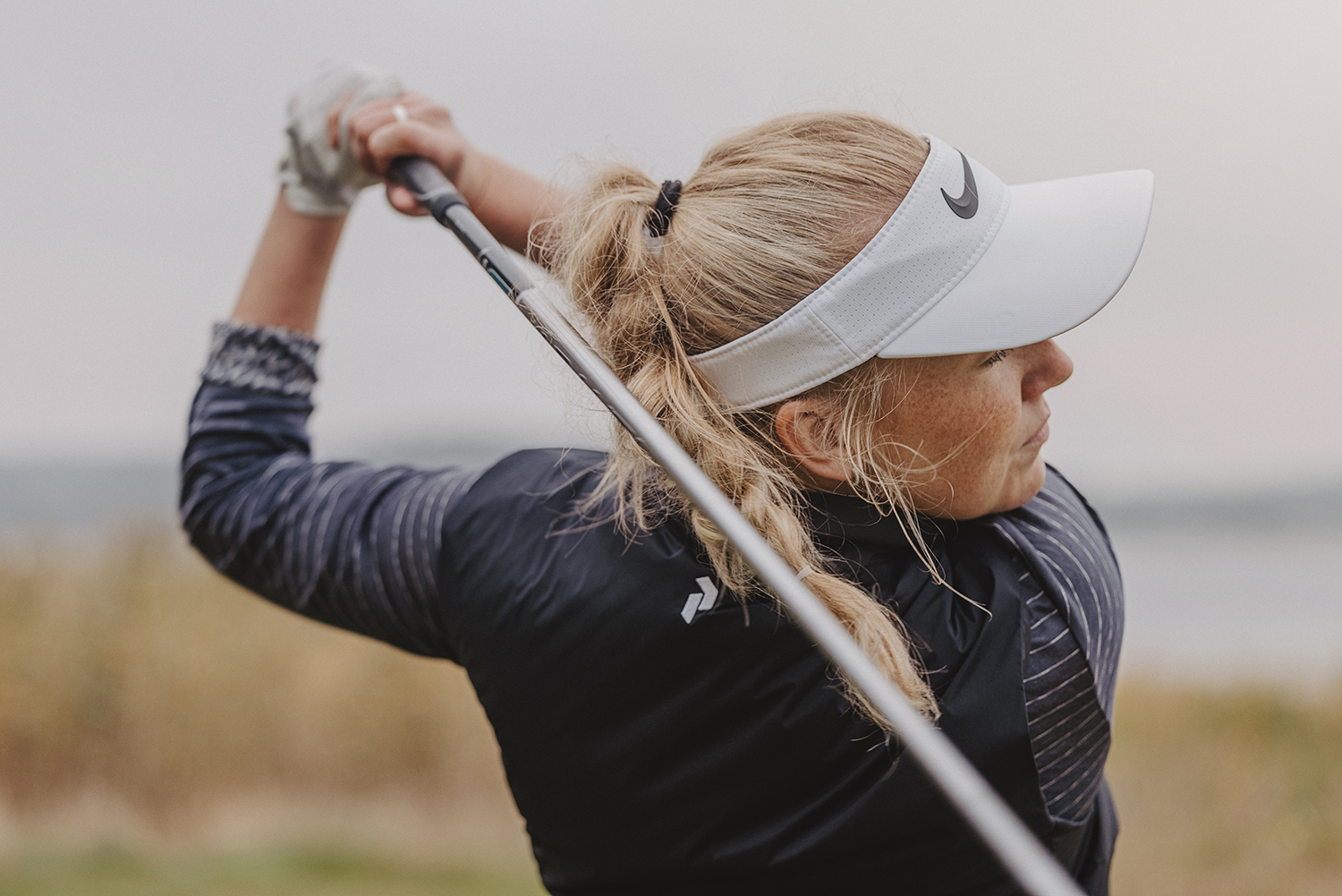Kvinna med blond hästsvans svingar en golfklubba utomhus, bär vit skärm med Nike-logga och mörk jacka.