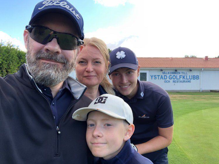 Fyra personer poserar tillsammans på en golfbana framför en byggnad med skylten "Välkommen till Ystad Golfklubb och restaurang".