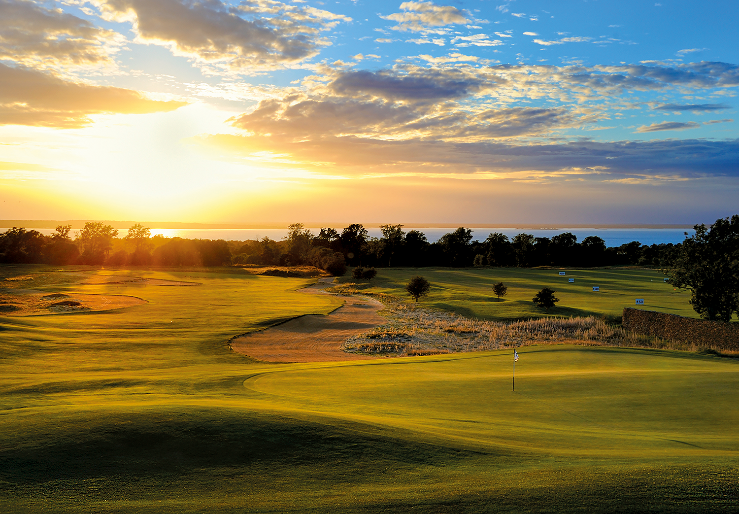 Golfbana med flagga på green, solnedgång i bakgrunden och träd längs horisonten.