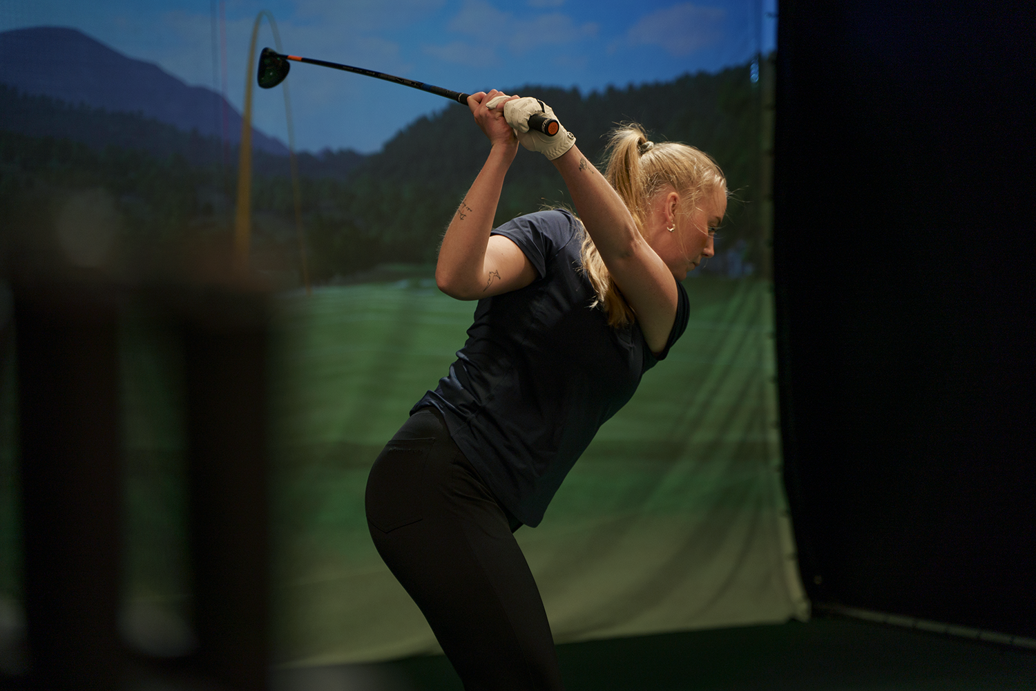 Kvinna med hästsvans svingar en golfklubba inomhus framför en stor skärm med golfbana i bakgrunden.