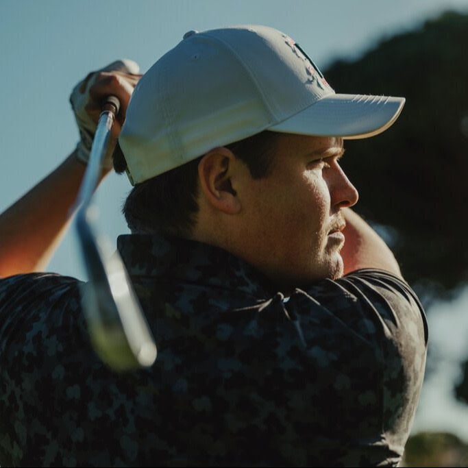 Man svingar en golfklubba utomhus, bär vit keps och mörk mönstrad tröja, med träd i bakgrunden.