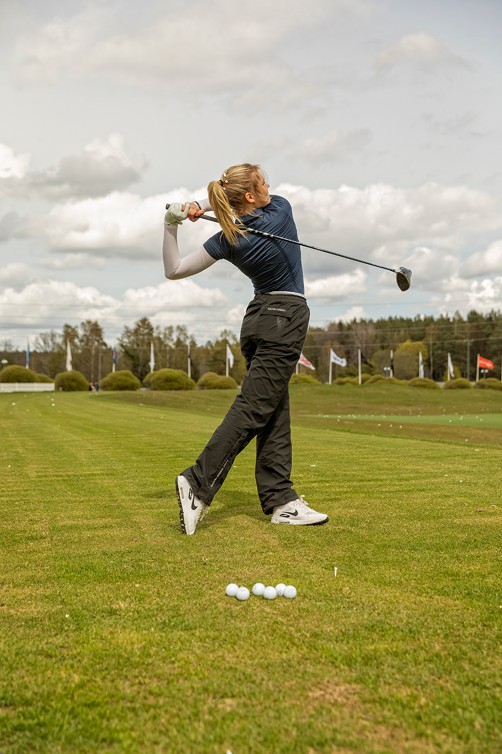 Kvinna svingar en golfklubba på driving range, golfbollar ligger på gräset framför henne, flaggor i bakgrunden.