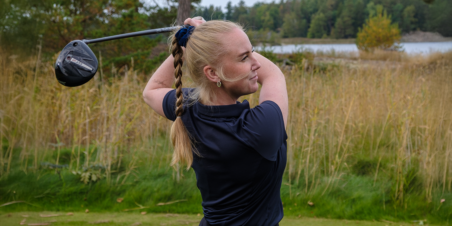 Kvinna med fläta svingar en golfklubba på en golfbana med högt gräs och träd i bakgrunden.