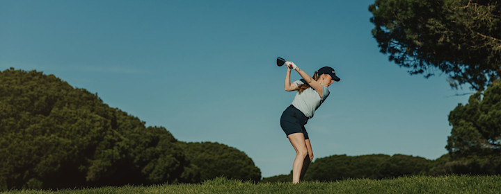 Kvinna i golfkläder svingar en golfklubba på en grön golfbana med träd och blå himmel i bakgrunden.