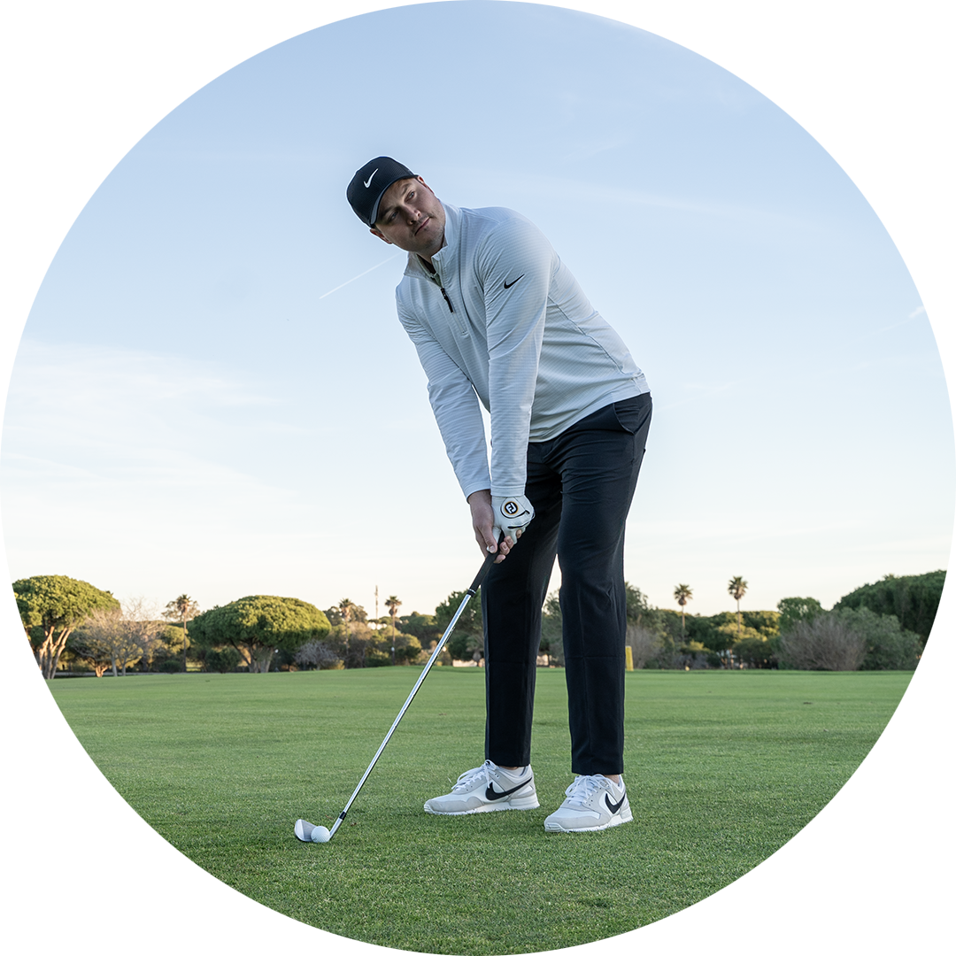 Man i sportkläder förbereder sig för att slå en golfboll på en grön golfbana med träd i bakgrunden.