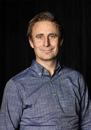 Man med kort brunt hår och blå skjorta står och ler mot kameran framför en mörk bakgrund.