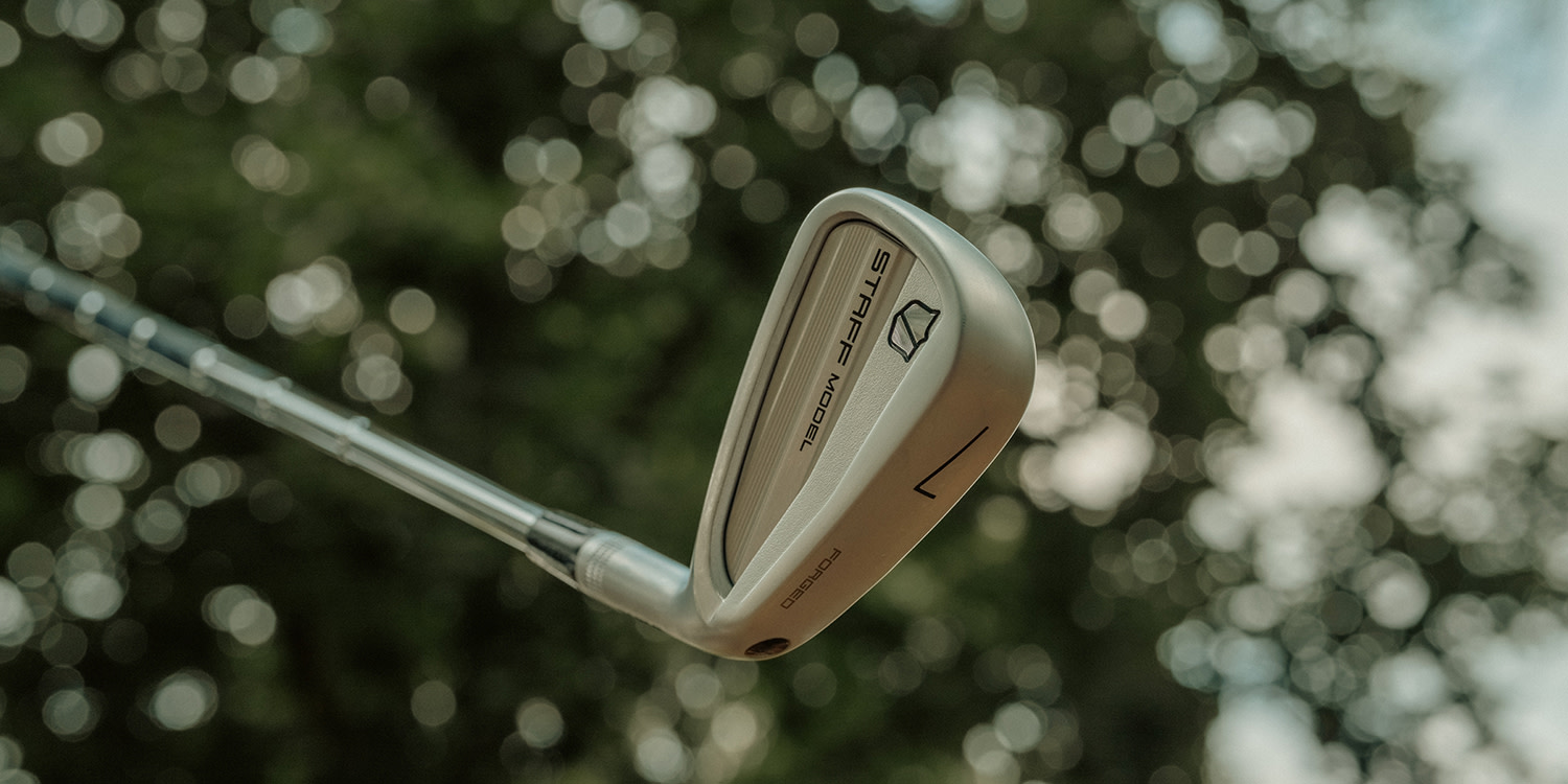 En golfklubba med texten "STAFF MODEL" hålls upp mot en suddig bakgrund av gröna träd och ljusprickar.