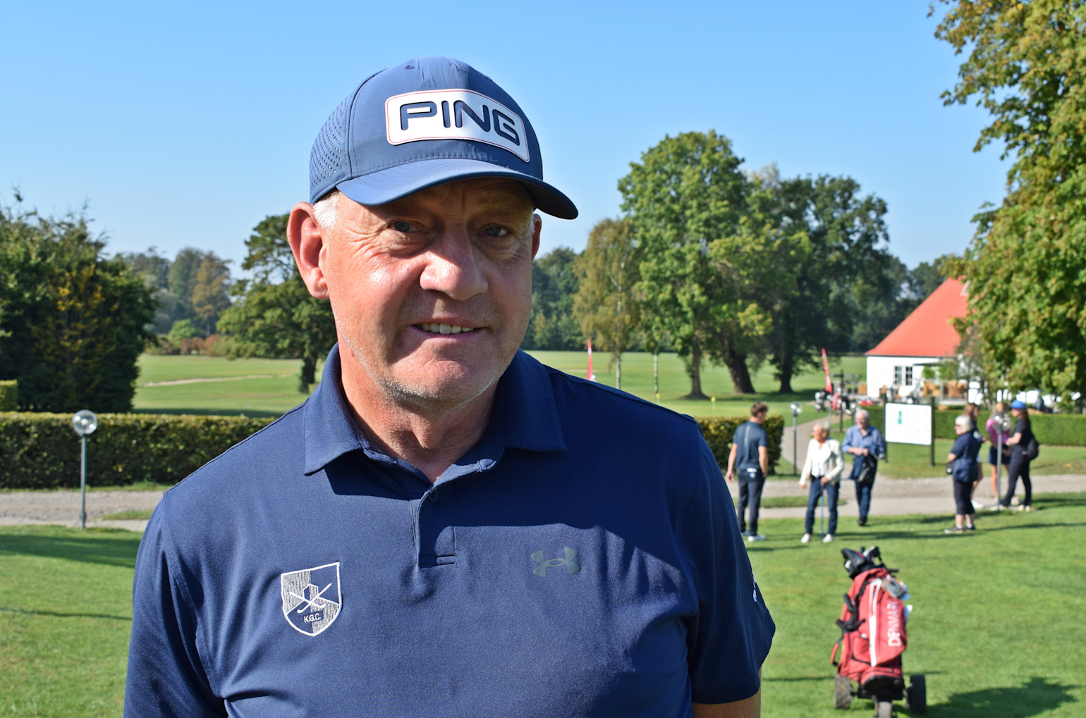 Man med blå PING-keps och golftröja står på en golfbana, med människor och klubbhus i bakgrunden under en solig dag.