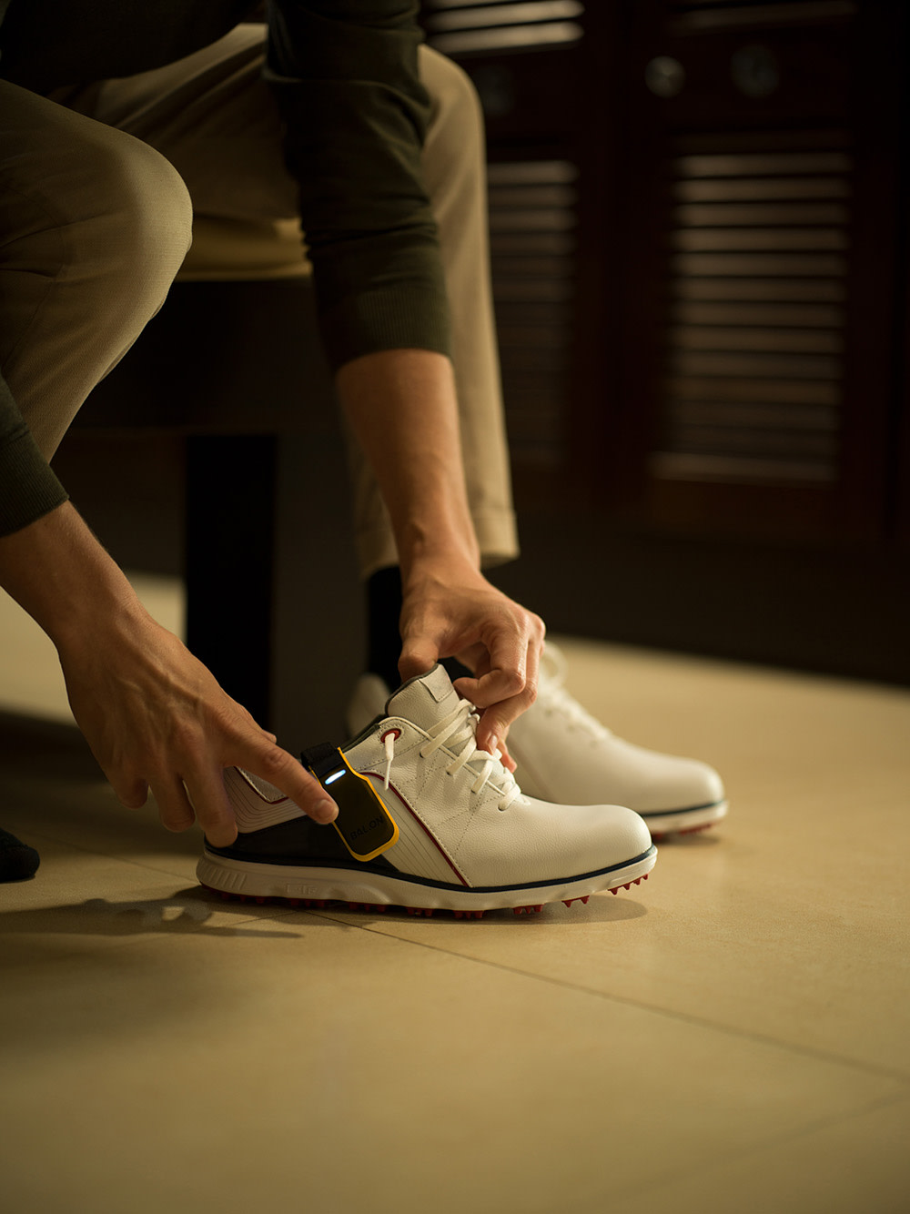 Person sätter på sig vita golfskor med svarta och röda detaljer, sittande på en bänk i ett omklädningsrum.