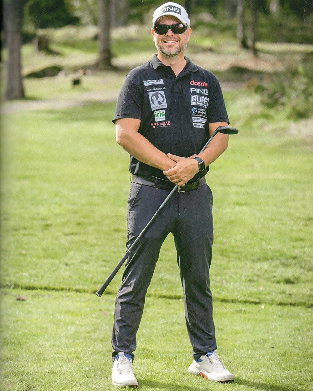 Man med solglasögon och keps står leende på en golfbana, håller en golfklubba och bär svart pikétröja med sponsormärken.