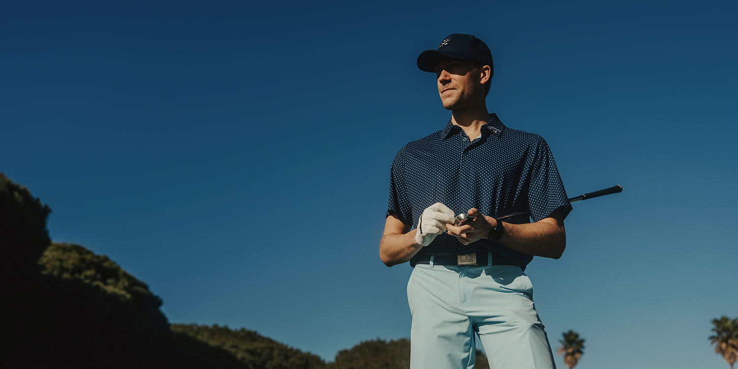 A man in golf attire, holding a golf glove and club, stands outdoors on a golf course under a clear blue sky.