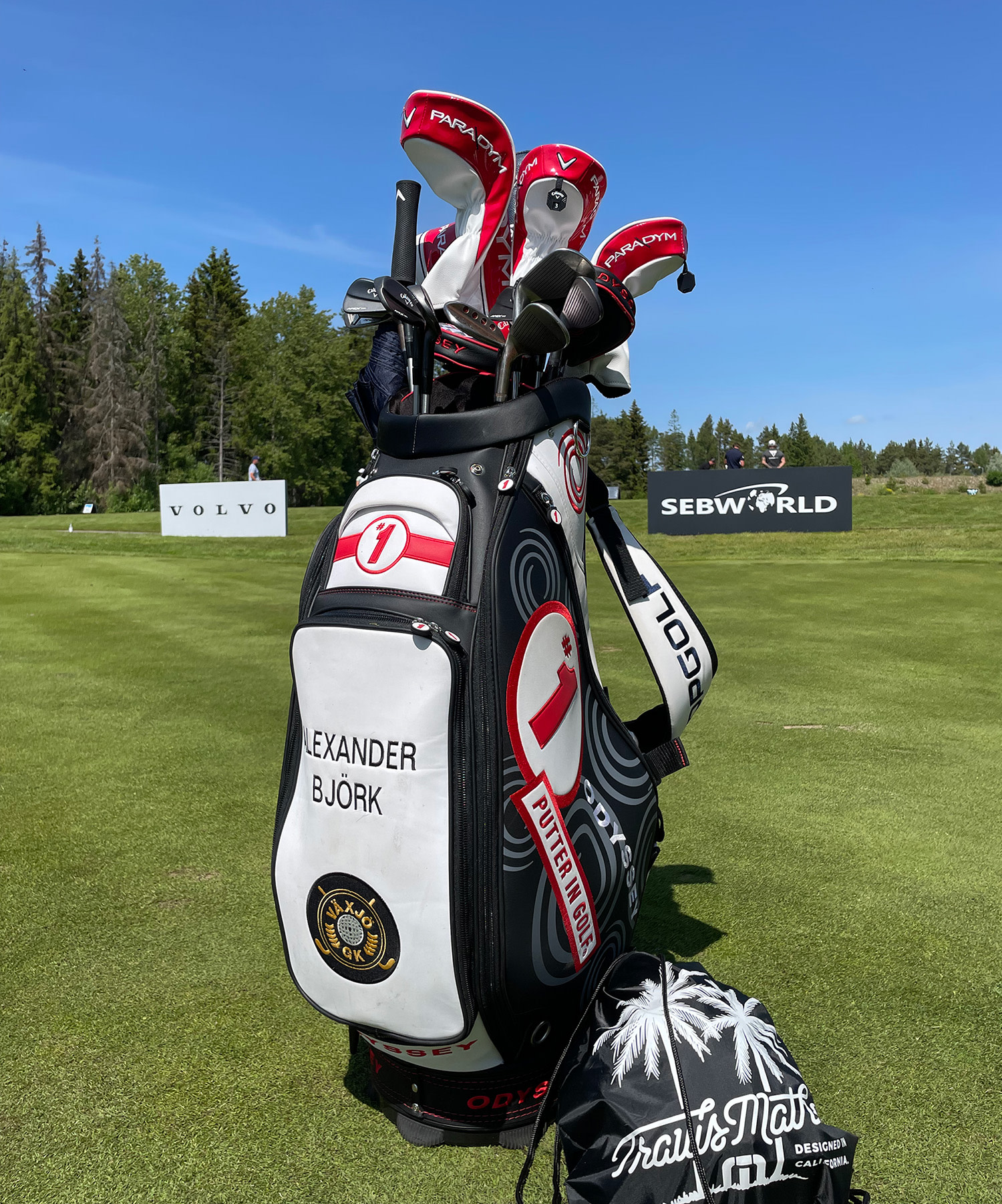 Golfbag med texten "Alexander Björk" står på en golfbana, omgiven av klubbor och headcovers, med skyltar för Volvo och SEB World i bakgrunden.