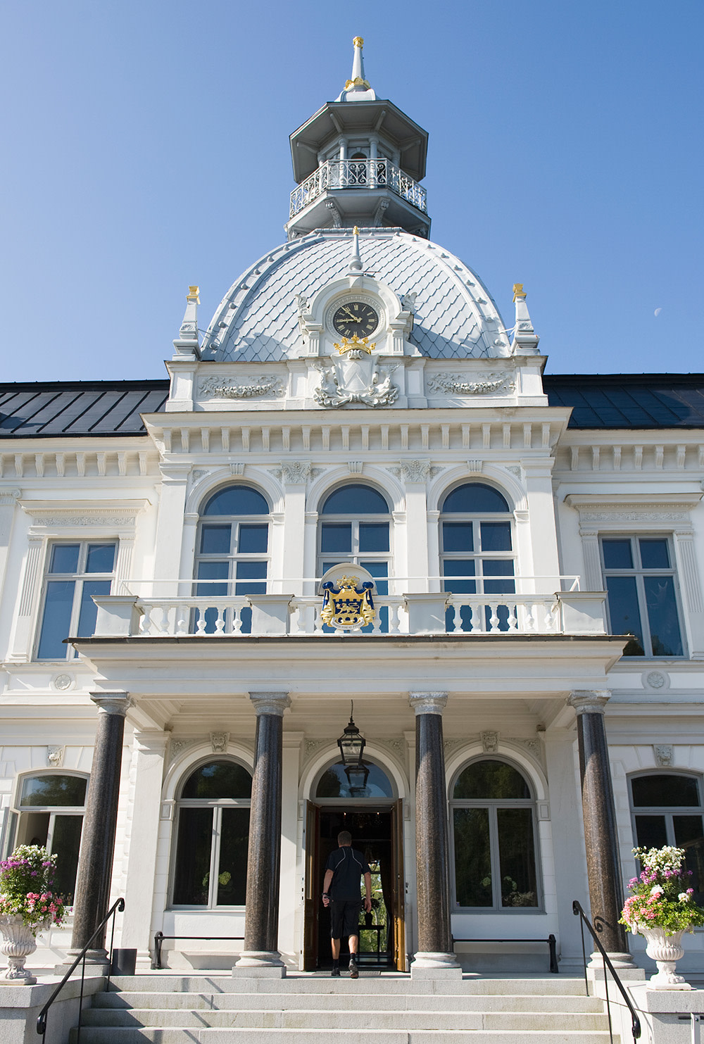 Vit herrgårdsbyggnad med kupol, klocka och gulddetaljer, person går in genom huvudingången, blomkrukor vid trappan, blå himmel.