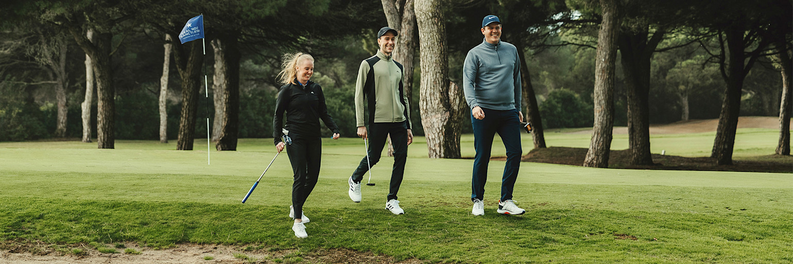 Tre personer går och ler på en golfbana med träd i bakgrunden, en golfklubba och flagga syns.