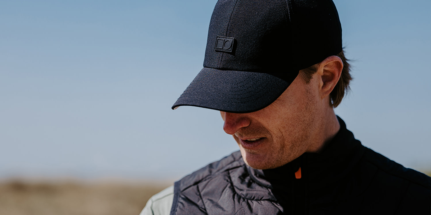 Man med svart keps och svart väst tittar ner utomhus i soligt väder, suddig bakgrund med blå himmel.