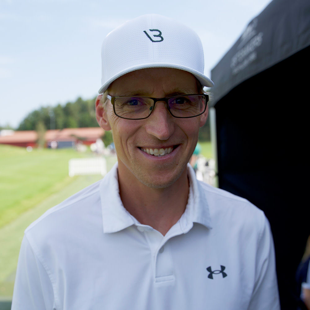 Man med glasögon och vit keps ler, klädd i vit pikétröja med Under Armour-logotyp, utomhus vid en golfbana.