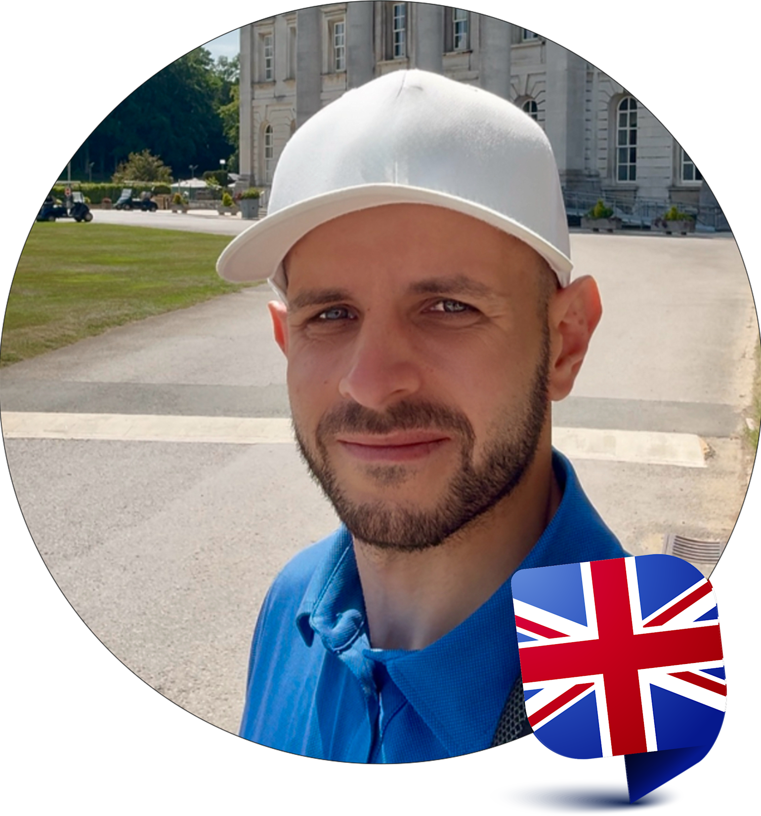Man med skägg och vit keps ler utomhus framför en byggnad, med brittisk flagga i nedre högra hörnet.