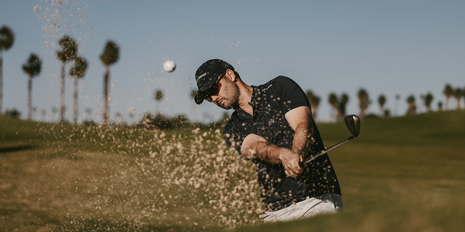 A man hits a golf ball out of a sand bunker on a golf course with palm trees in the background on a sunny day.