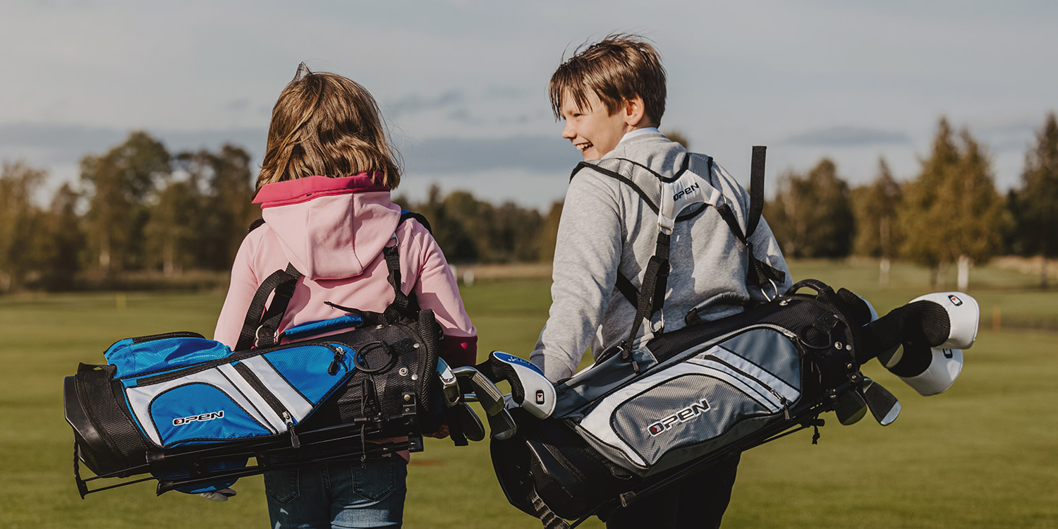 Två barn bär golfbagar och går tillsammans på en golfbana, omgivna av grönt gräs och träd i bakgrunden.