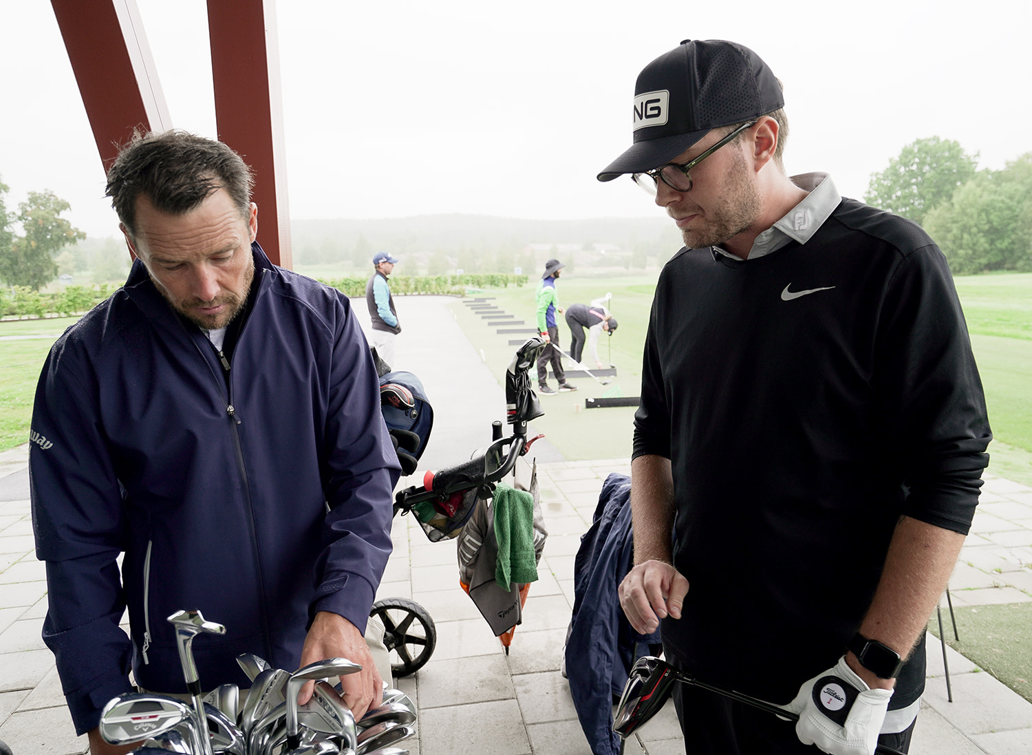 Två män står vid en golfbana, en sorterar golfklubbor medan den andra håller en klubba, med fler personer som tränar i bakgrunden.