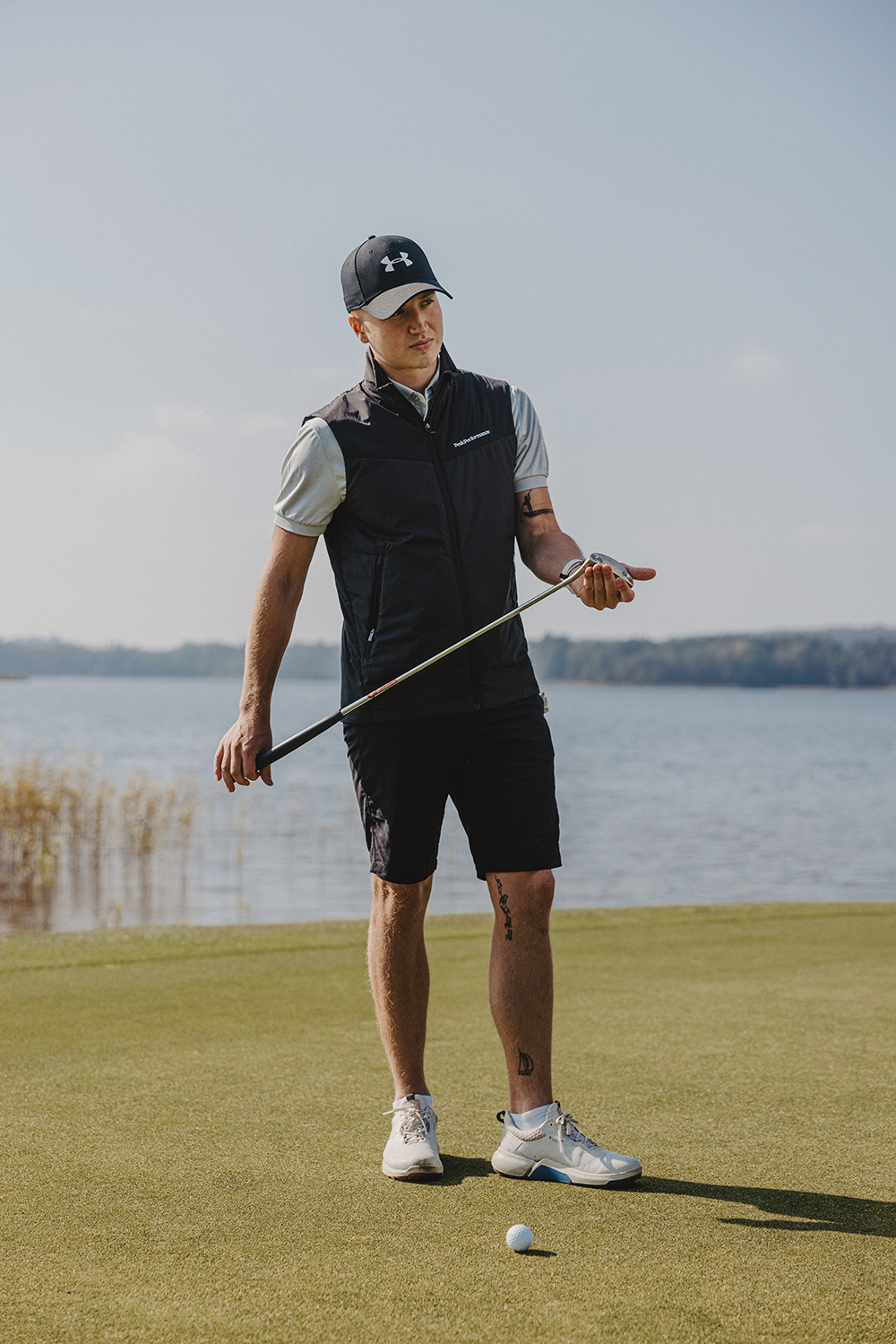 Man i golfkläder håller en golfklubba och står på en green nära en sjö, med en golfboll framför sig.