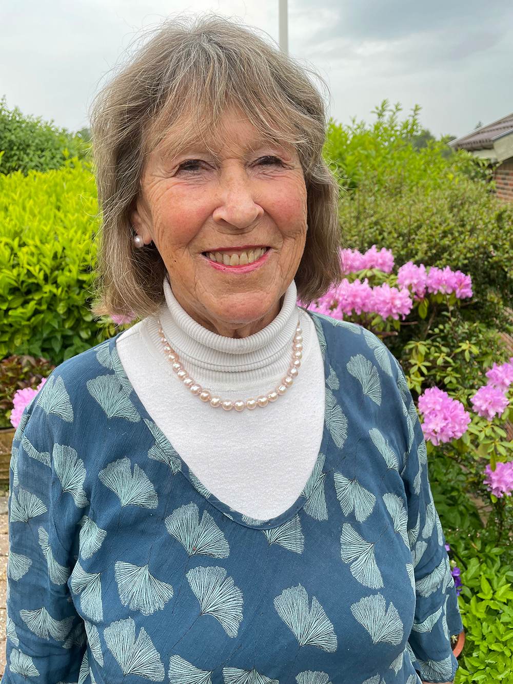 Äldre kvinna med grått hår och pärlhalsband ler utomhus, omgiven av gröna buskar och rosa blommor.