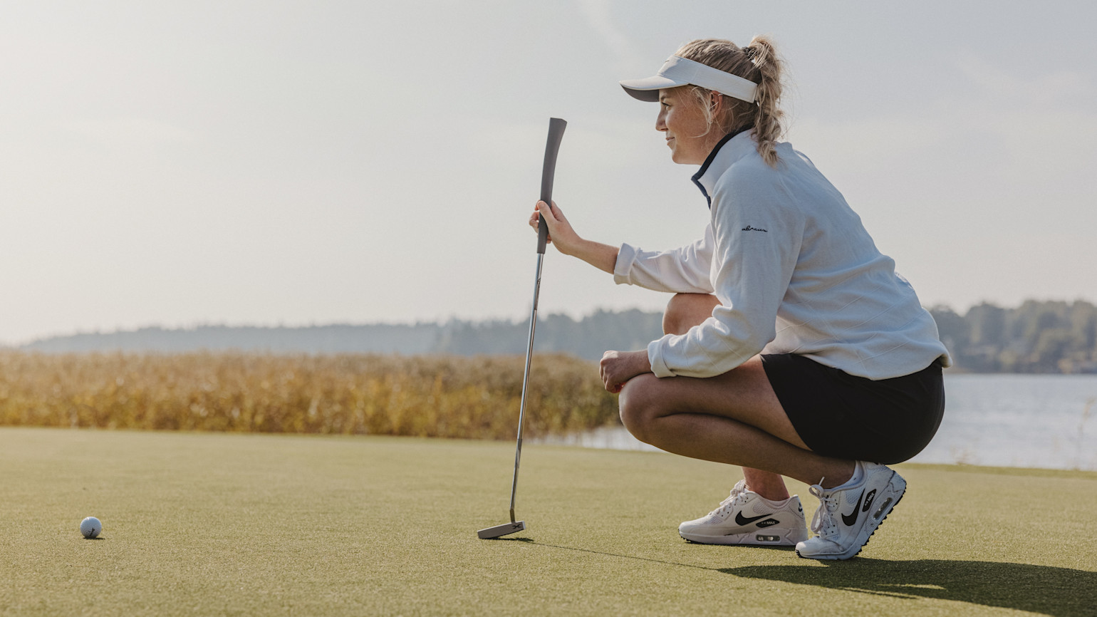 A woman in a white top and visor crouches with a golf club, aiming at a golf ball on the green by the water.