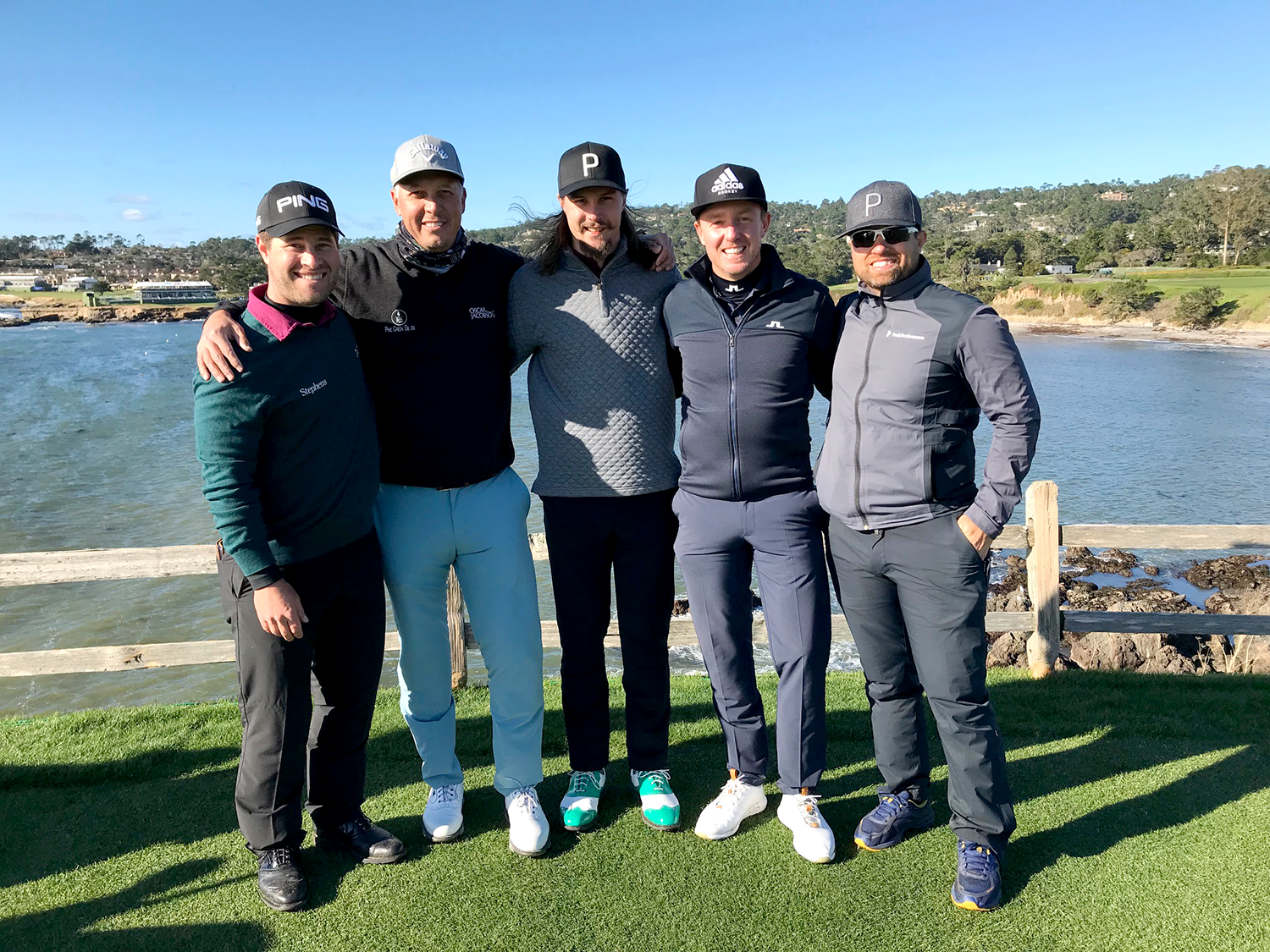 Fem män står tillsammans på en golfbana vid vattnet, klädda i golfkläder och kepsar, med armarna om varandra under en solig dag.
