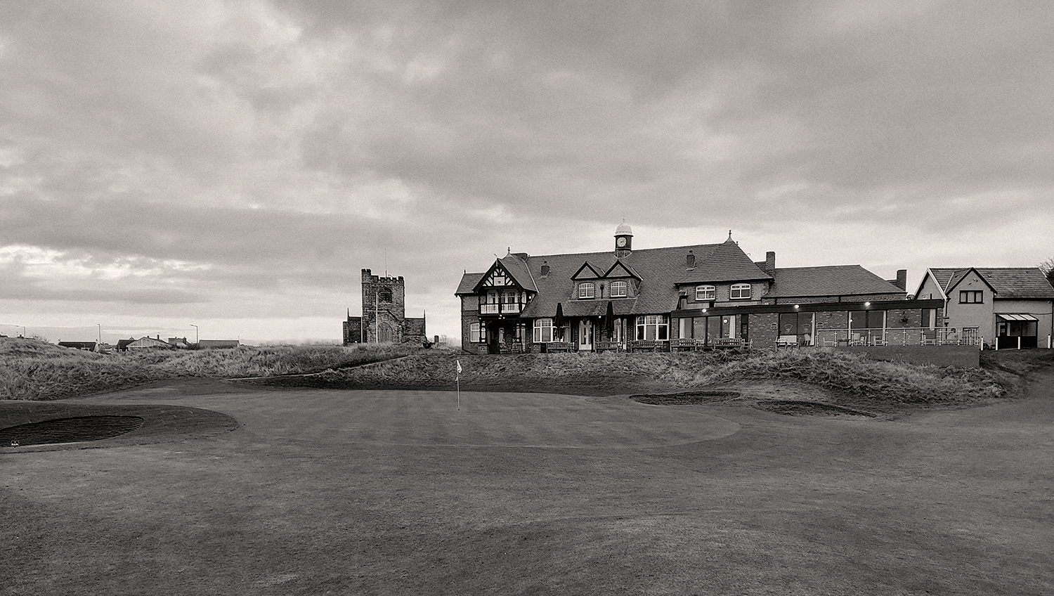 En golfgreen med flagga i förgrunden, klubbhus och gammal stenkyrka i bakgrunden under molnig himmel.