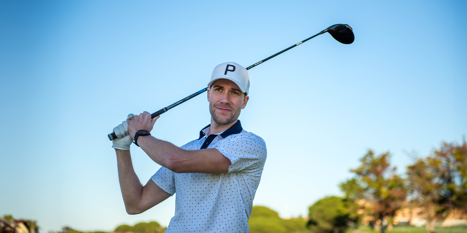 Man i vit keps med bokstaven P håller en golfklubba över axlarna på en golfbana under klarblå himmel.