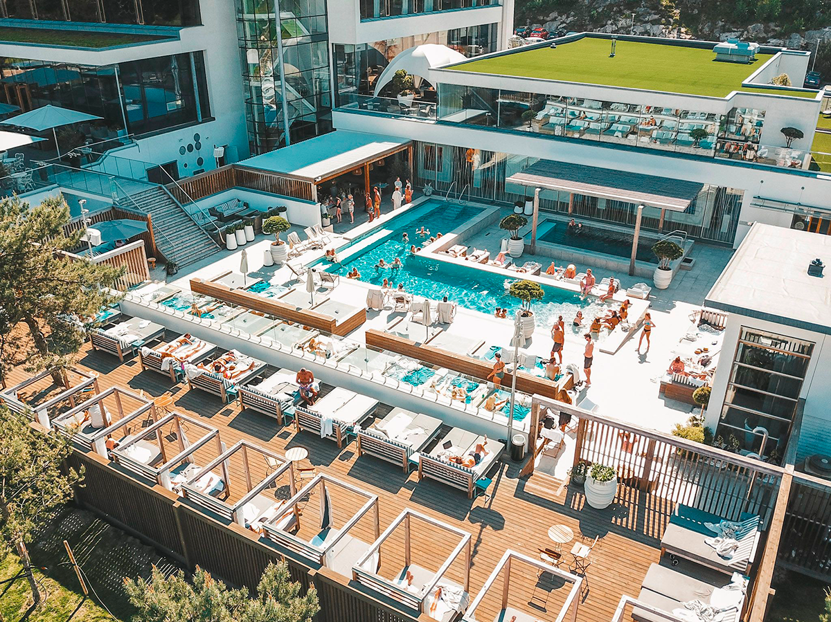 Människor solar och badar vid en modern utomhuspool på ett lyxigt hotell med solbäddar och cabanas runt poolområdet.