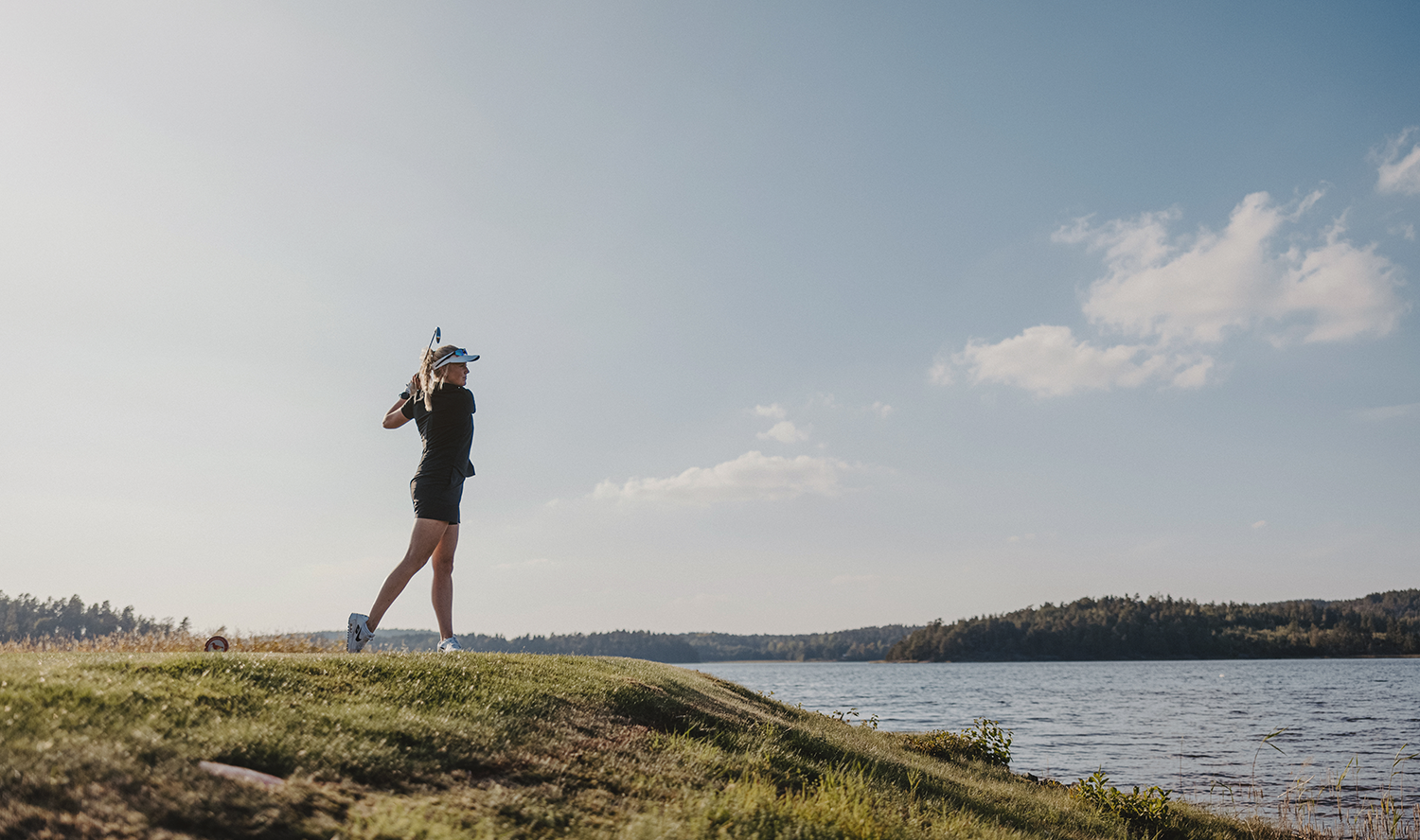 Kvinna svingar en golfklubba på en grön golfbana vid vattnet under en klarblå himmel.