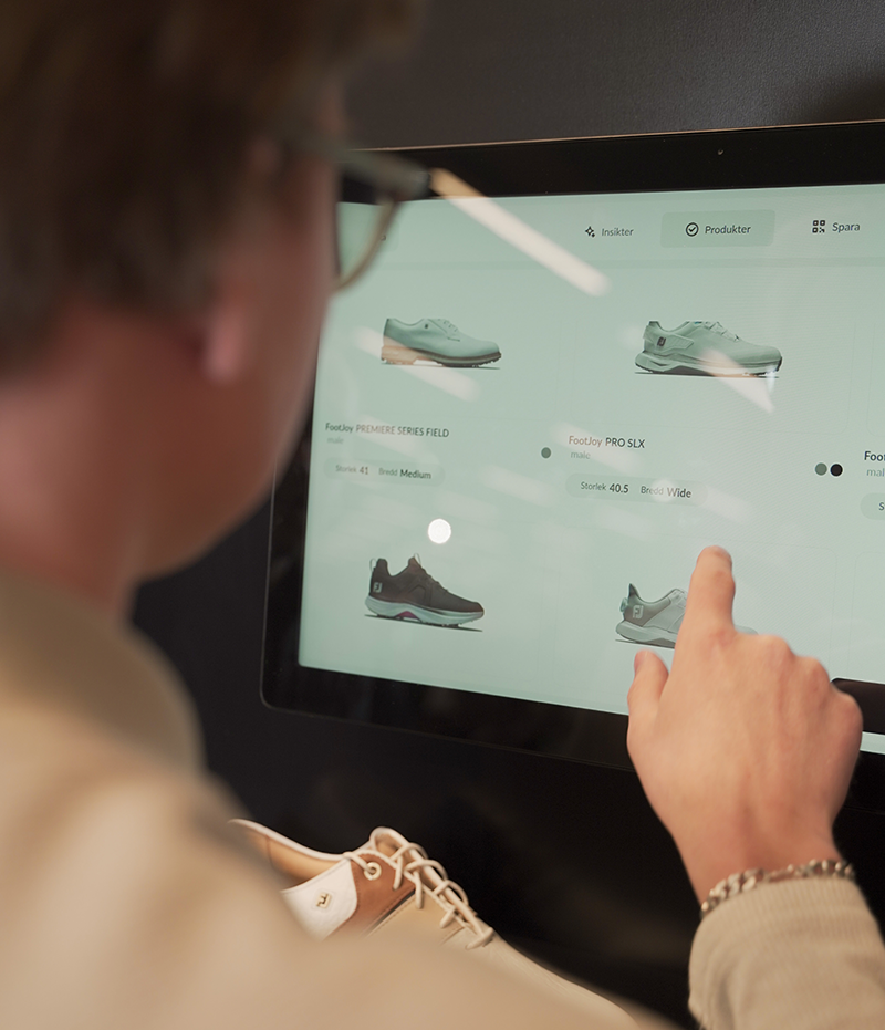 Person pekar på en pekskärm och bläddrar bland olika skor i en digital butiksmiljö.