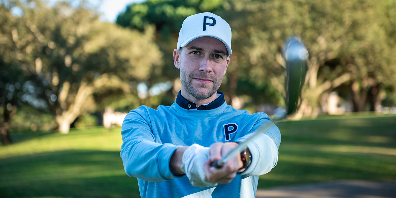 Man in a blue golf shirt and a white cap with the letter P is holding a golf club in front of him on a green golf course.
