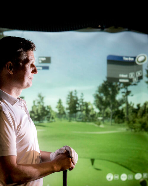 Man i vit pikétröja håller en golfklubba och tittar på en golfsimulator med en virtuell golfbana i bakgrunden.