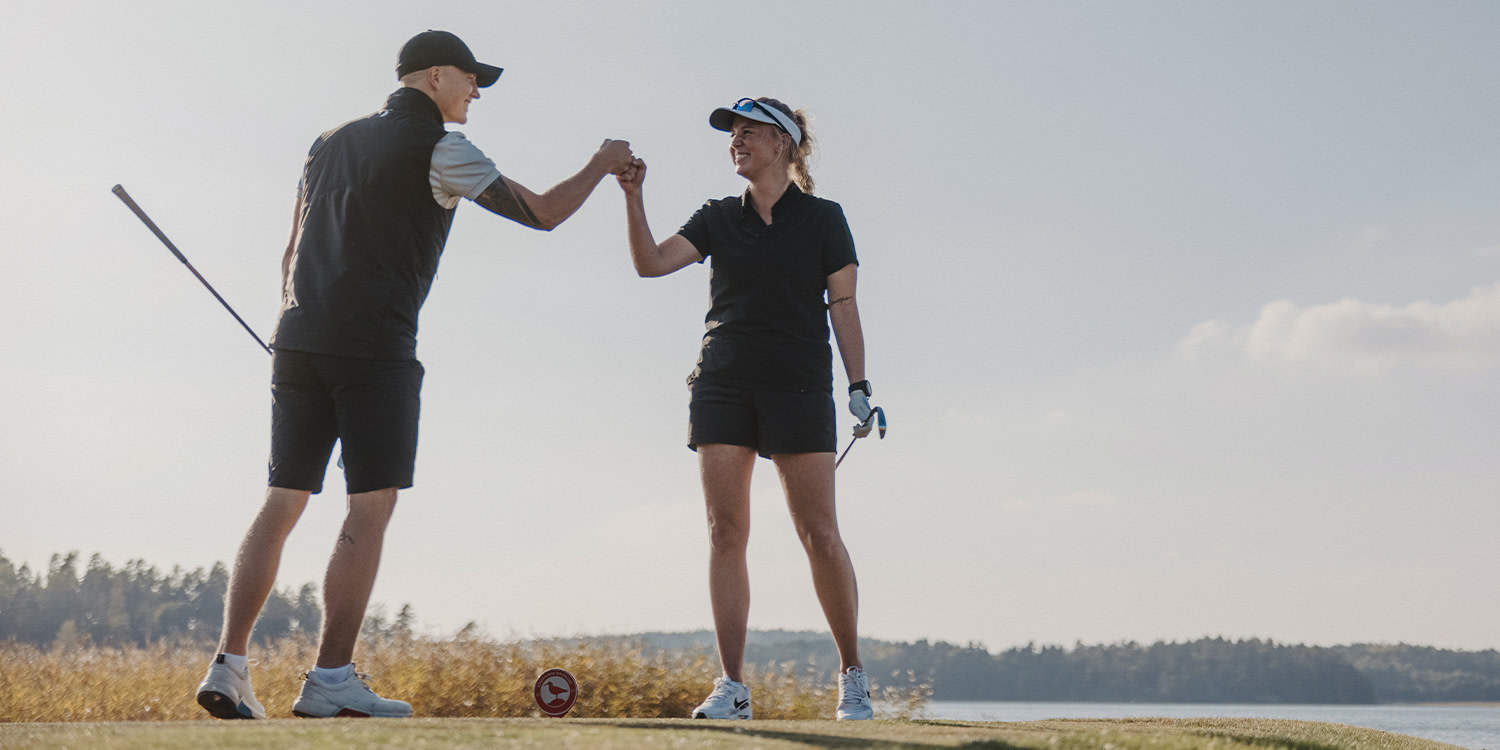Two golfers give each other a fist bump on the golf course, both holding golf clubs and smiling at each other with a lake in the background.