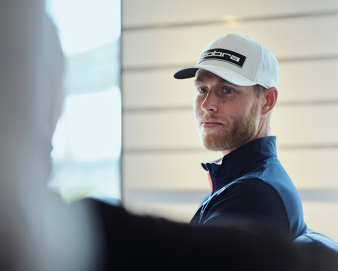 Man med skägg och vit keps med texten "Cobra" sitter inomhus och tittar åt sidan, klädd i mörkblå jacka.
