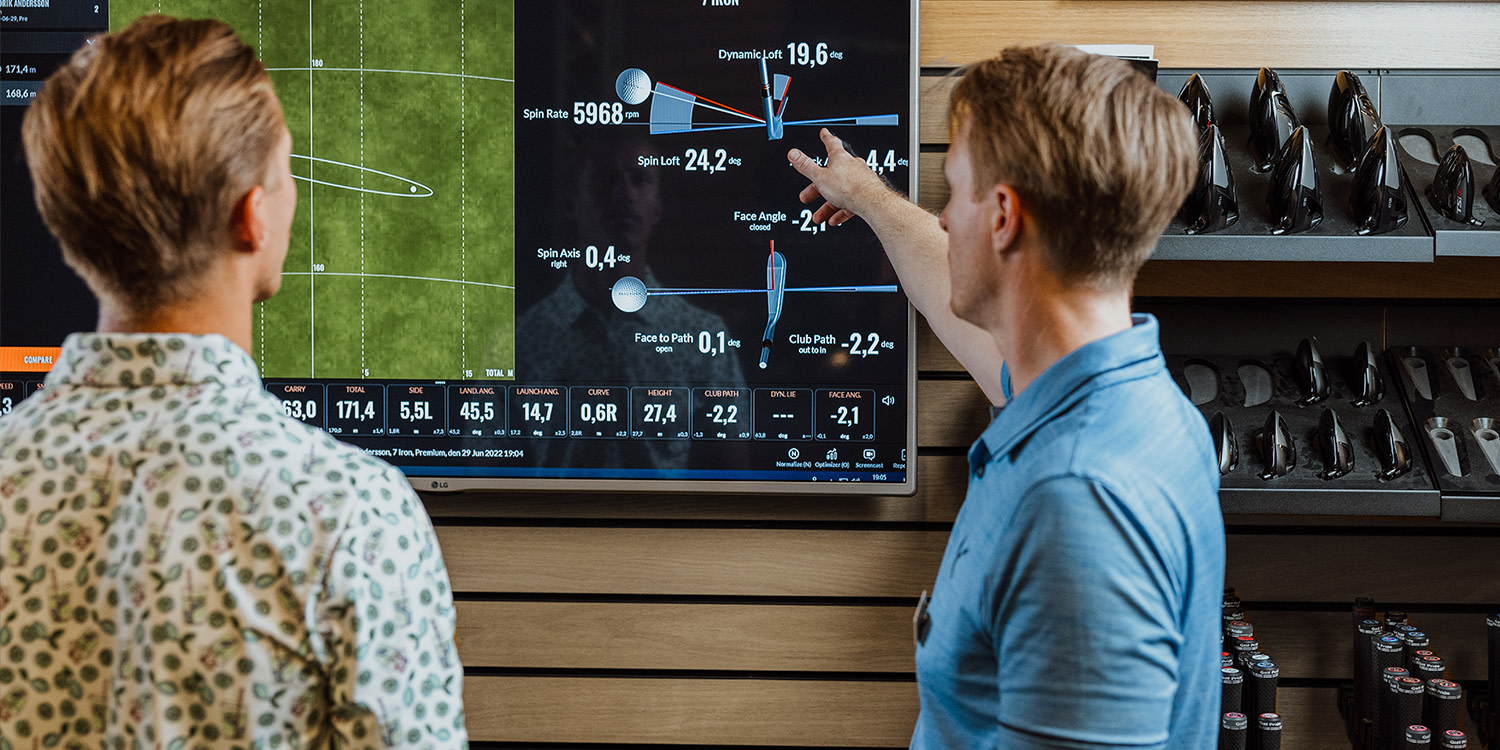 Två män analyserar golfdata på en stor skärm, en pekar på siffror och grafer i en golfbutiksmiljö.