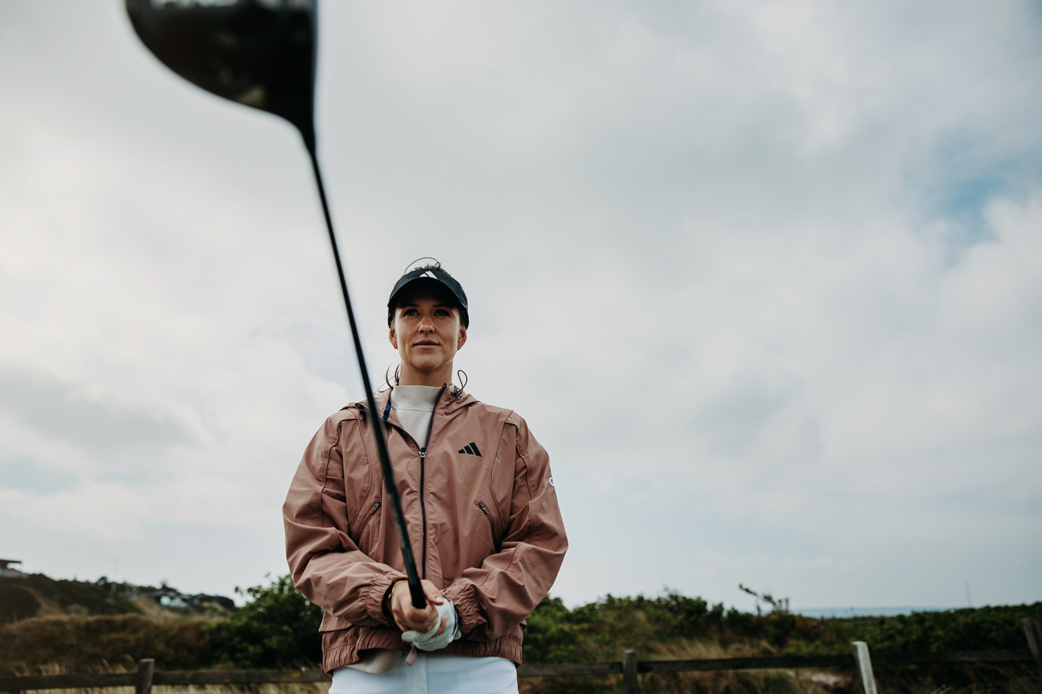 Kvinna i sportjacka och keps håller en golfklubba utsträckt mot kameran på en golfbana under molnig himmel.