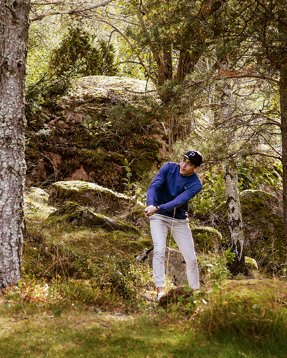Man i blå tröja och keps slår ett golfslag från skogsterräng omgiven av träd och mossa.