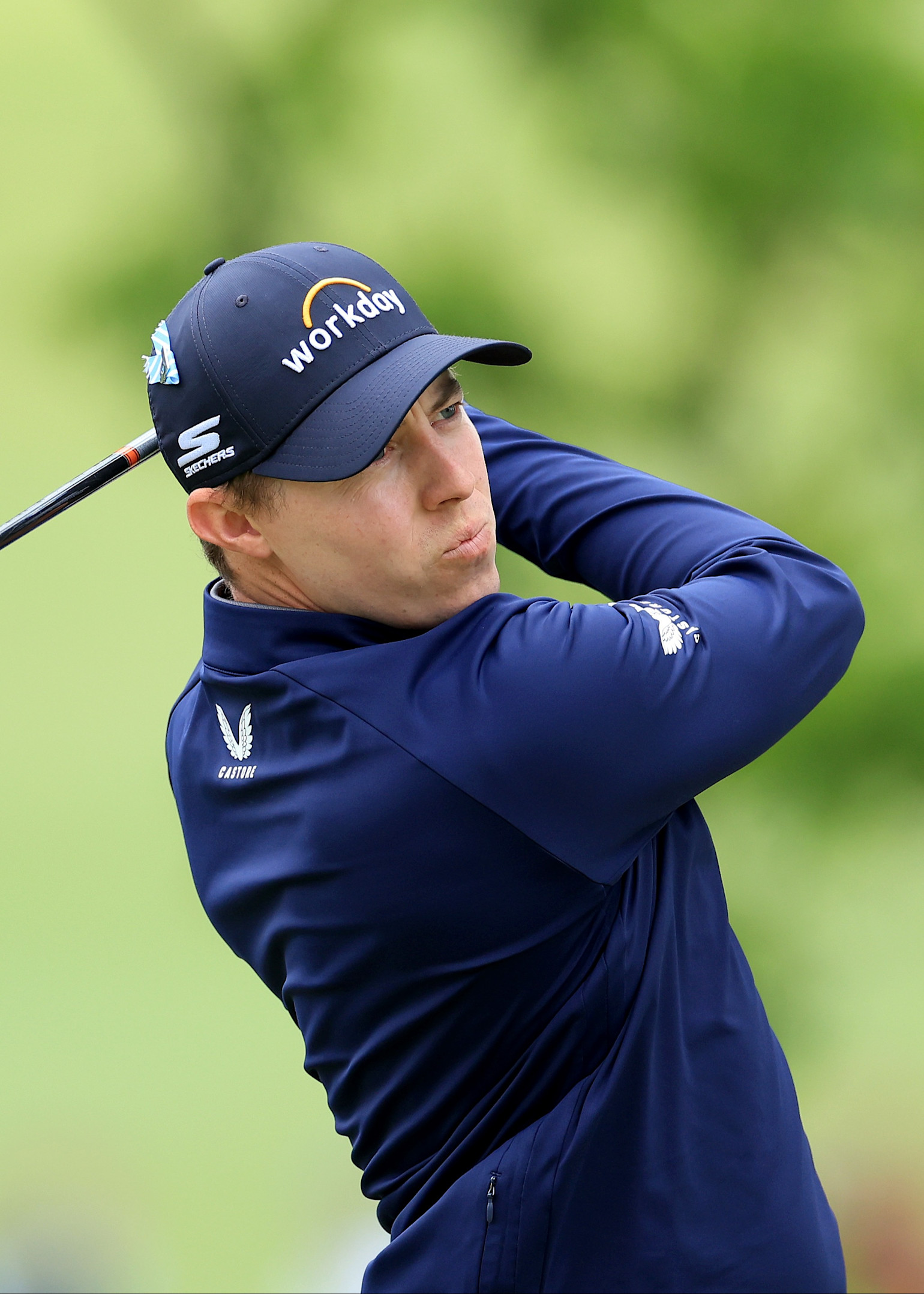 Golfspelare svingar en klubba på en golfbana, bär blå keps med texten "workday" och blå jacka.