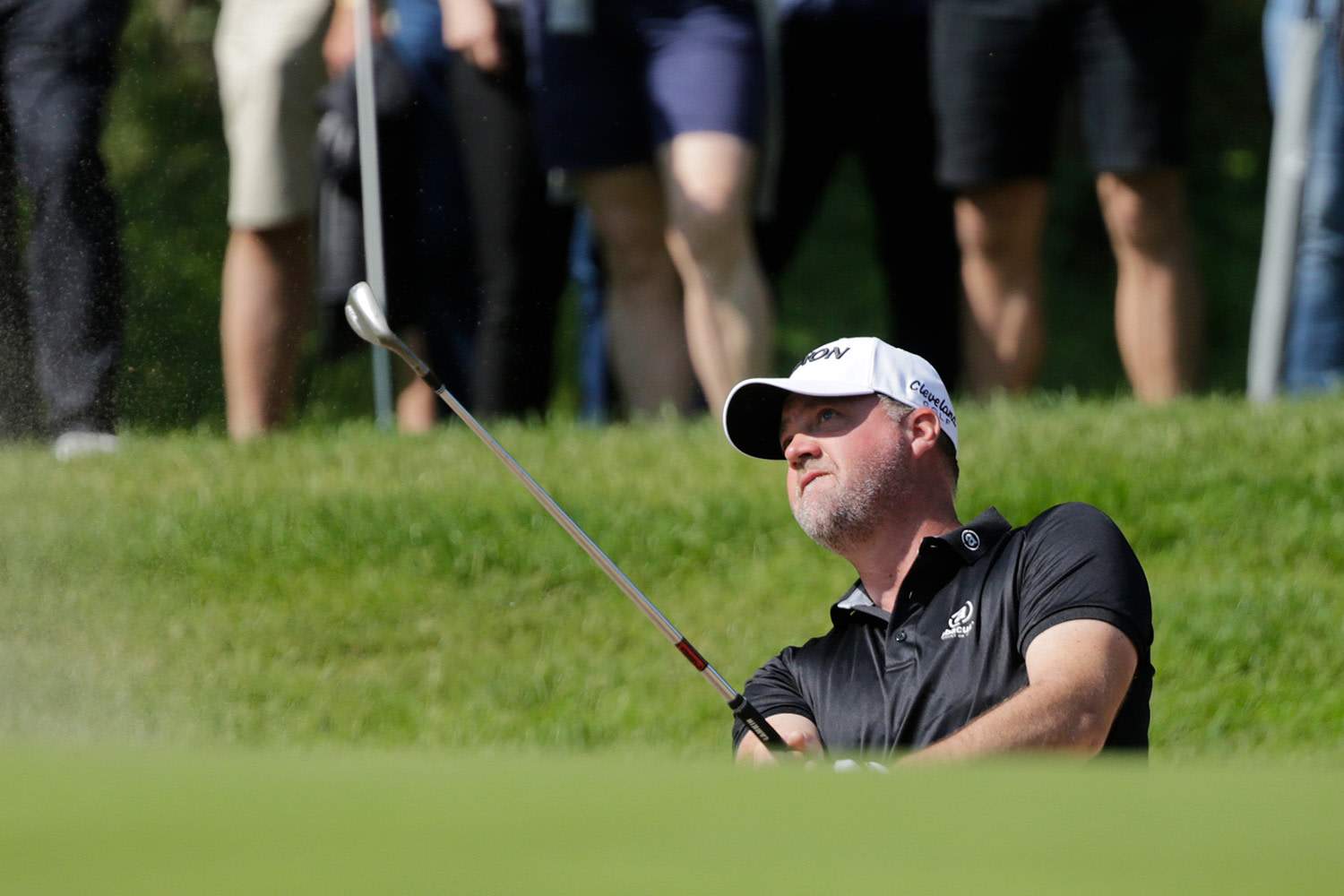 Golfspelare slår ett slag ur en bunker med en sandwedge, åskådare står i bakgrunden på gräset.