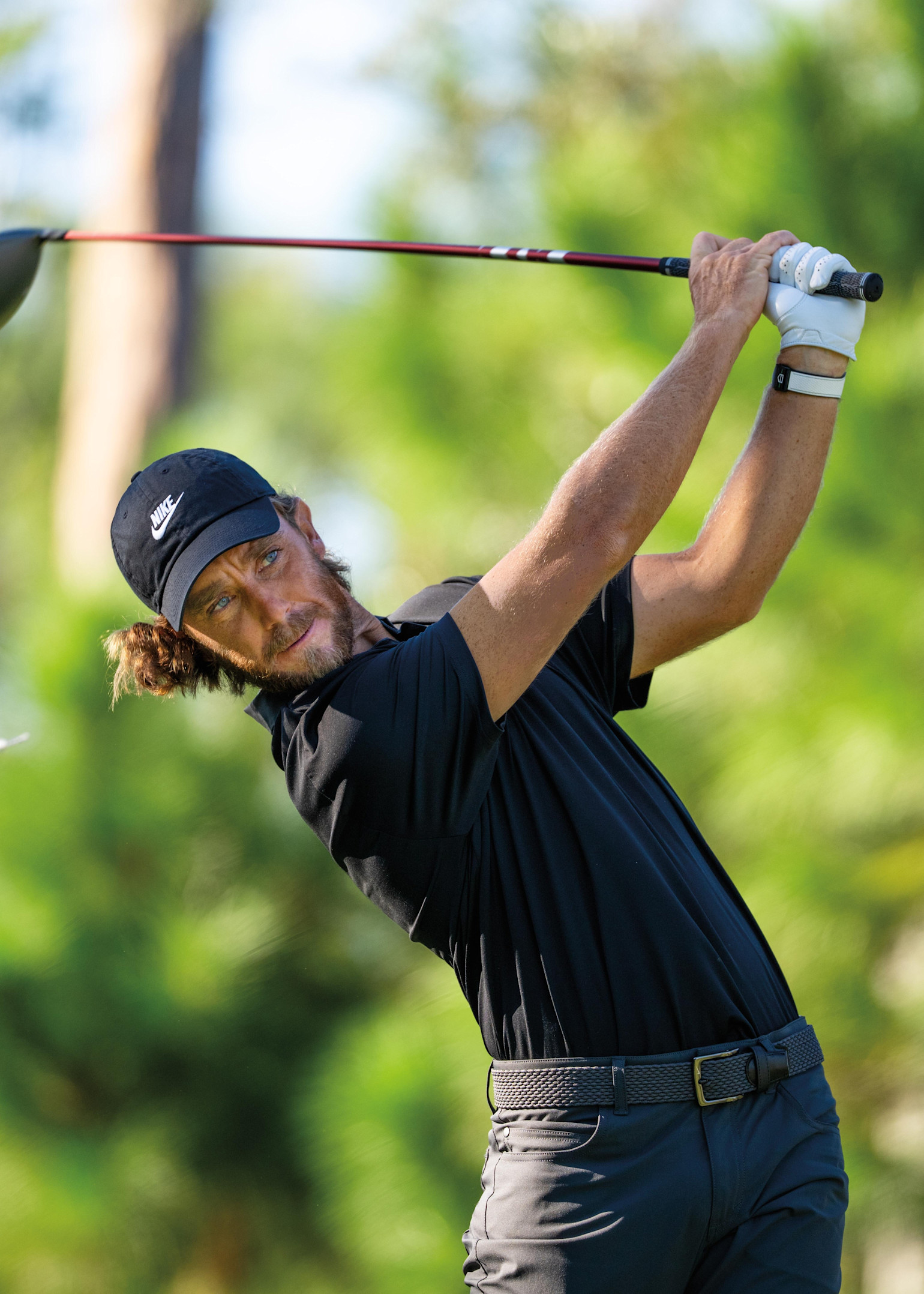 Man i svart pikétröja och keps svingar en golfklubba på en golfbana med grönskande bakgrund.