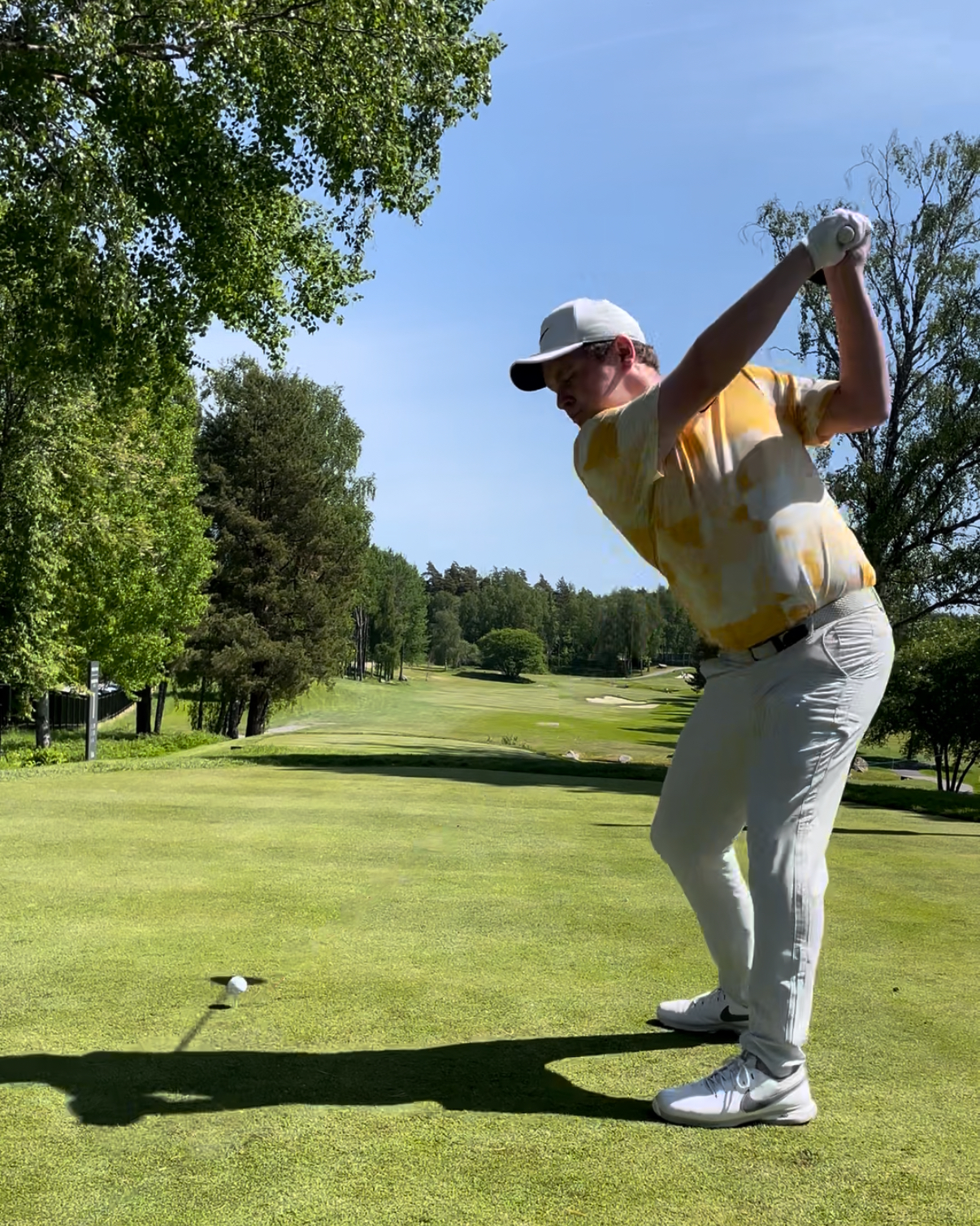 En golfspelare i gulvit skjorta och vit keps svingar en klubba på en grön golfbana omgiven av träd under klarblå himmel.