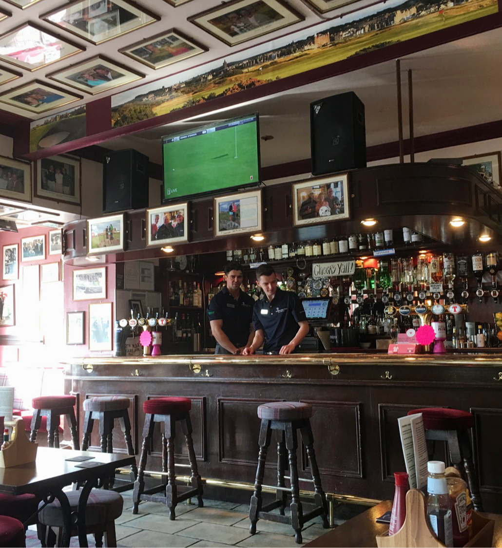 Två bartendrar arbetar bakom en träbar i en pub med många flaskor och kranar, väggar täckta av foton och en TV som visar sport.