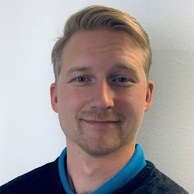 Man med blont hår och kort skägg ler mot kameran, står framför en vit vägg och bär mörk tröja med blå krage.