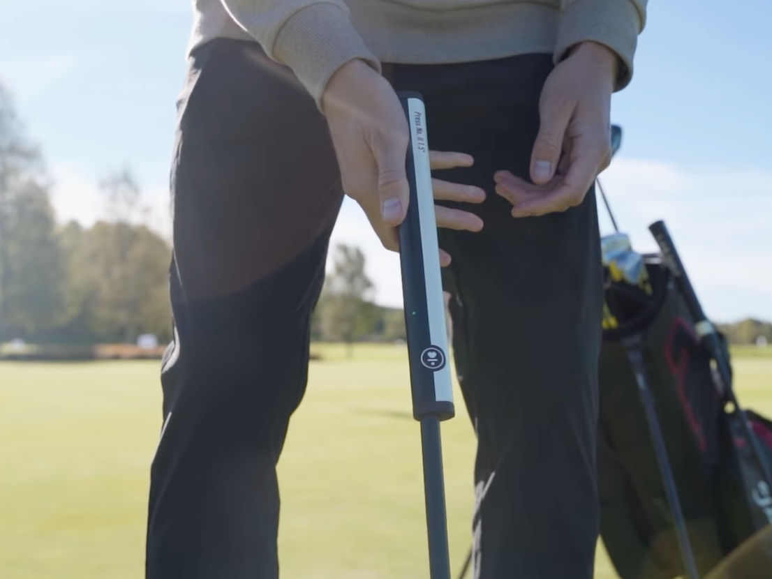 En person håller i ett golfklubbskaft på en golfbana, med en golfbag i bakgrunden under en solig himmel.